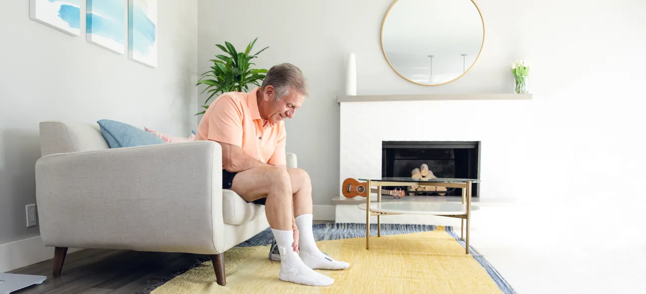 Elderly man sitting on a sofa in a living room, holding his ankle.